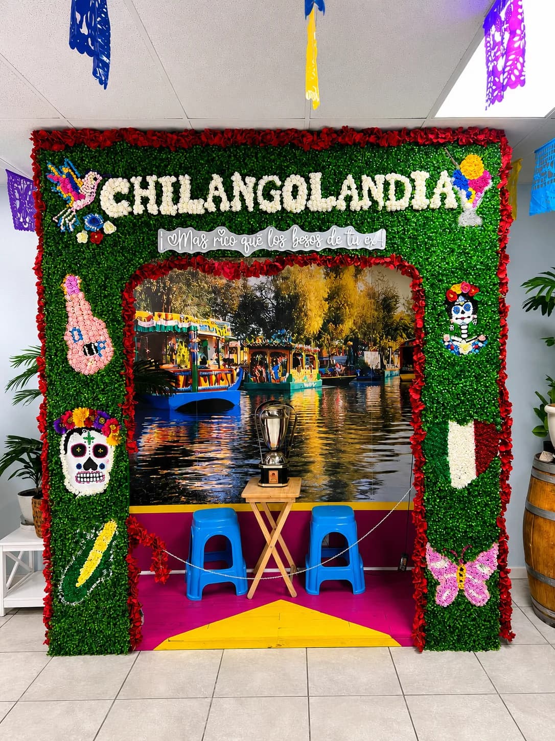Chilangolandia's floral photo wall spelling CHILANGOLANDIA in white blooms, framed with red flowers, sugar-skull art and a Xochimilco trajinera backdrop.