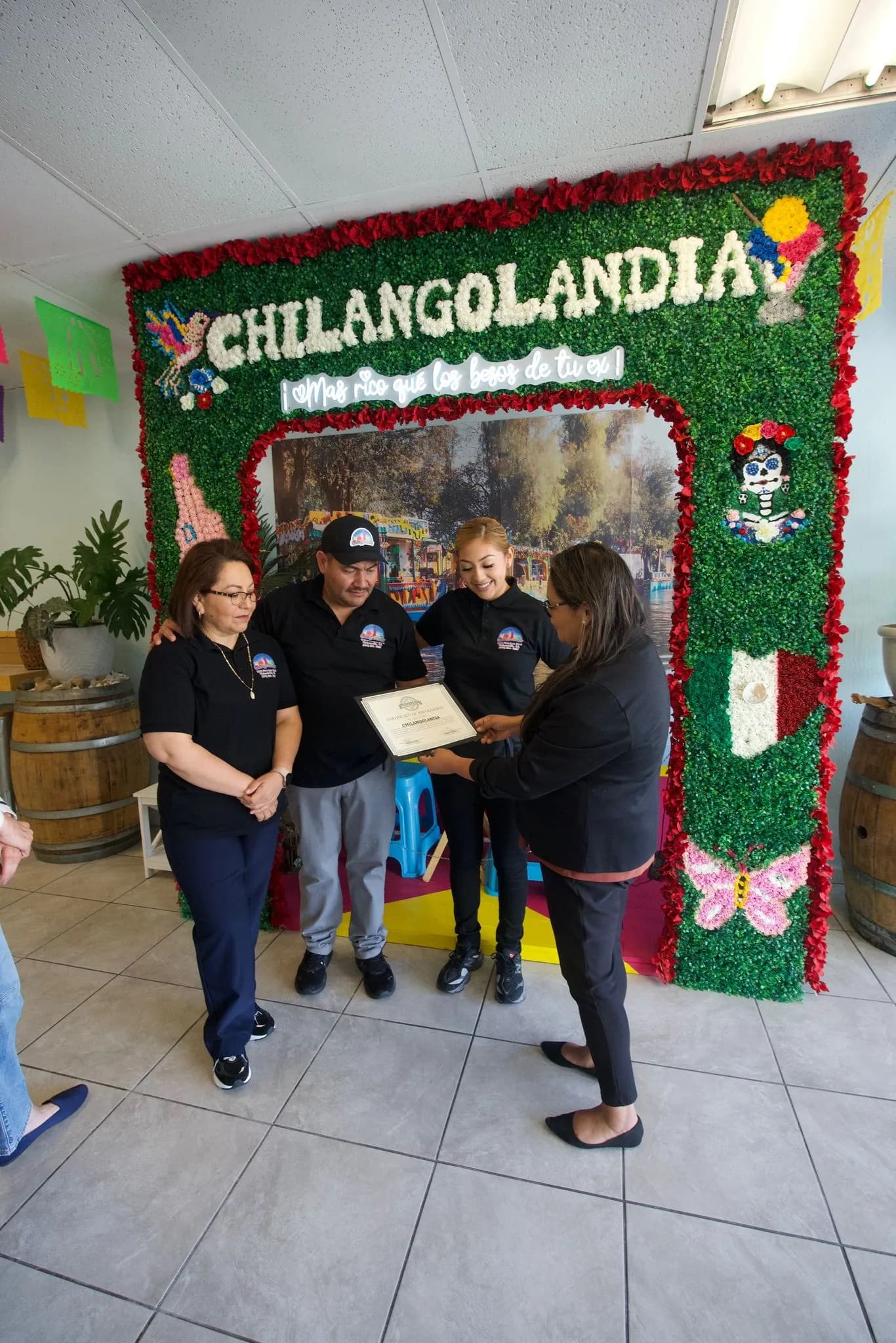 Chilangolandia staff in branded polo shirts holding a certificate in front of the floral photo wall.